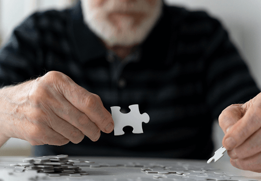 An older man holds a puzzle piece, looking thoughtfully at it, surrounded by scattered puzzle pieces on a table