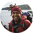 A smiling man with dreadlocks wearing a backpack stands against a neutral background.