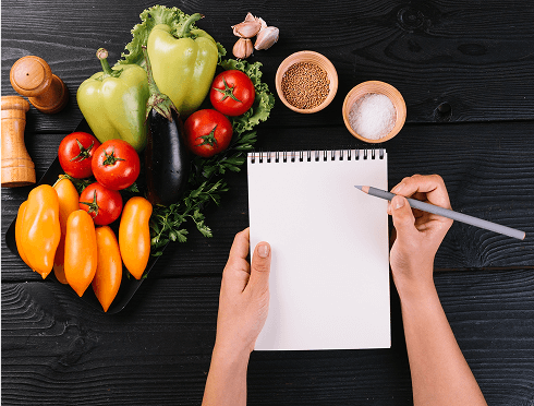 A person is jotting notes on a notepad with a pen, while fresh vegetables are arranged around them.